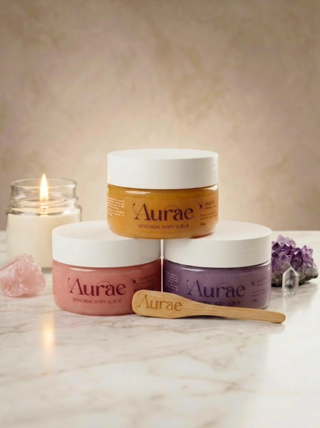 Os três potes do Sensorial Body Scrub da Aurae empilhados sobre mármore, nos aromas Âmbar, Quartzo Rosa e Ametista, com a espátula de madeira da marca, uma vela acesa e cristais de quartzo rosa e ametista ao lado